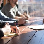 A group of people sit around a wooden table, taking notes during a collaborative meeting on the InterFAX vs Etherfax comparison.